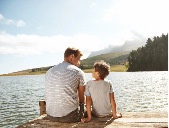 Father and son sitting together on a wooden dock by the water, sharing a quiet conversation.