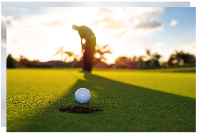Golf ball resting on the edge of the hole with a golfer putting in the background at sunset.