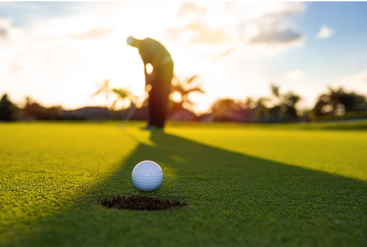 Golf ball near the hole on a putting green at sunset, with a golfer preparing to putt in the background.