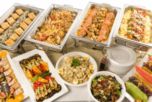 Buffet table with trays of pasta, salmon, chicken, vegetables, salad, and desserts.